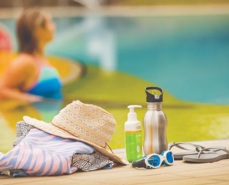 Clothing, sunscreen, swim goggles and a water bottle shown in focus by a pool