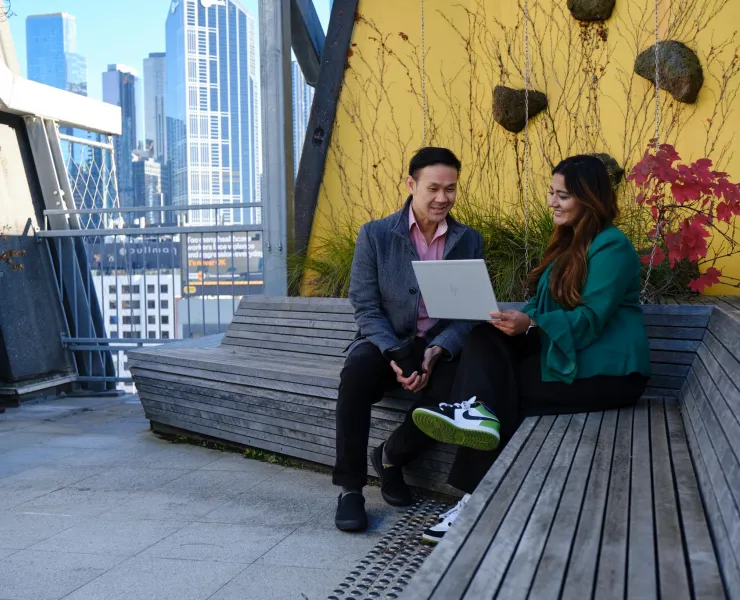 Two people sitting on a wooden bench looking at a laptop, with a prominent yellow painted wall with rocks hanging from it and a small shot of Melbourne's skyline.