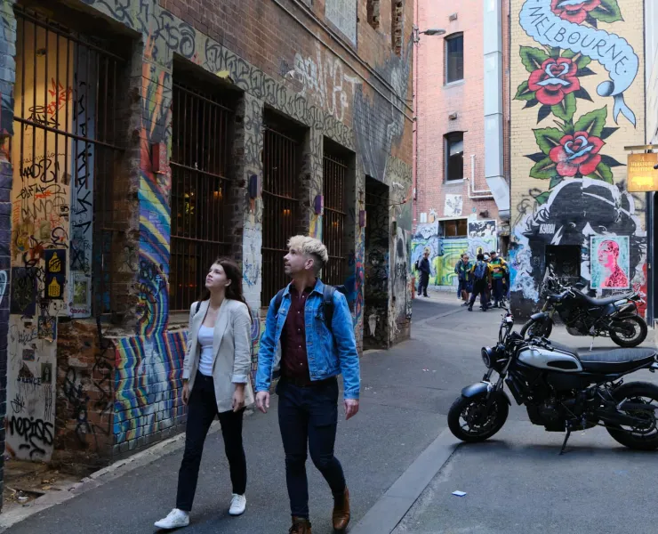 Walking through ACDC Lane, a couple passes by tourists gathered next to parked scooters, with iconic Melbourne graffiti art of roses adorning the building behind them.