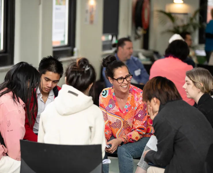 A group of six people sit together in a circle and appear to be engaging in a conversation with each other.