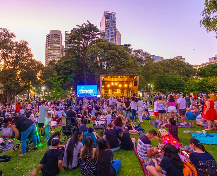 An audience seated on the lawn for a screen and stage event in a park.