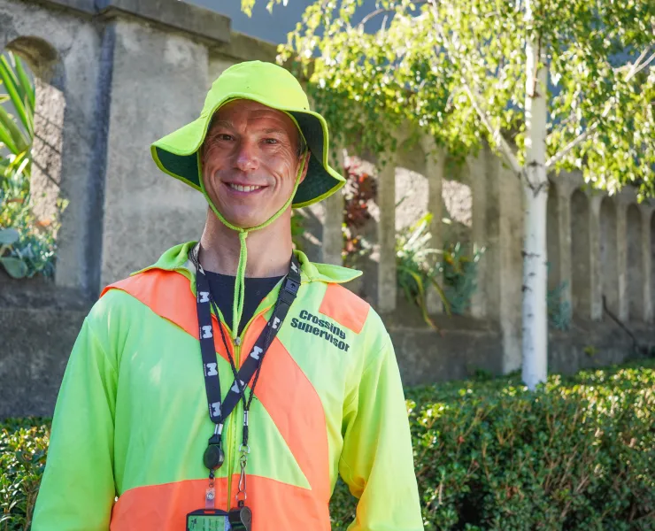 A school crossing supervisor wears a fluro jacket and bucket hat and a City of Melbourne lanyard. He is standing outside and smiling at the camera.