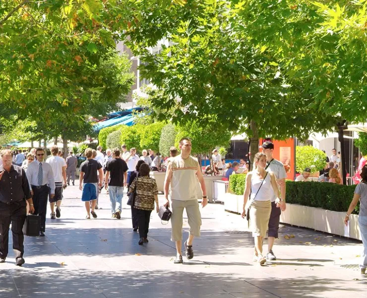 Lots of people walking along a wide promenade. There are cafes and trees alongside