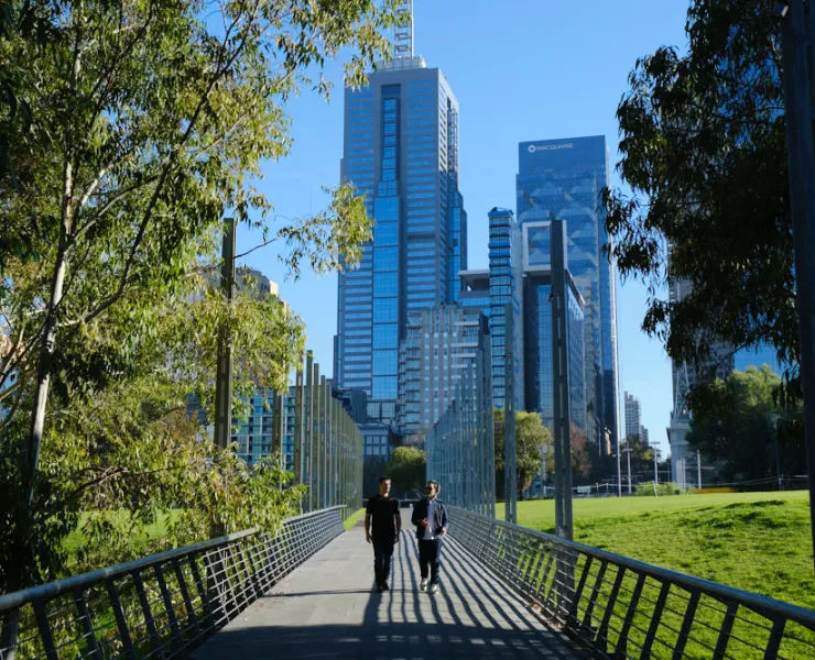 Two men walking down a walkway with tree and city buildings behind