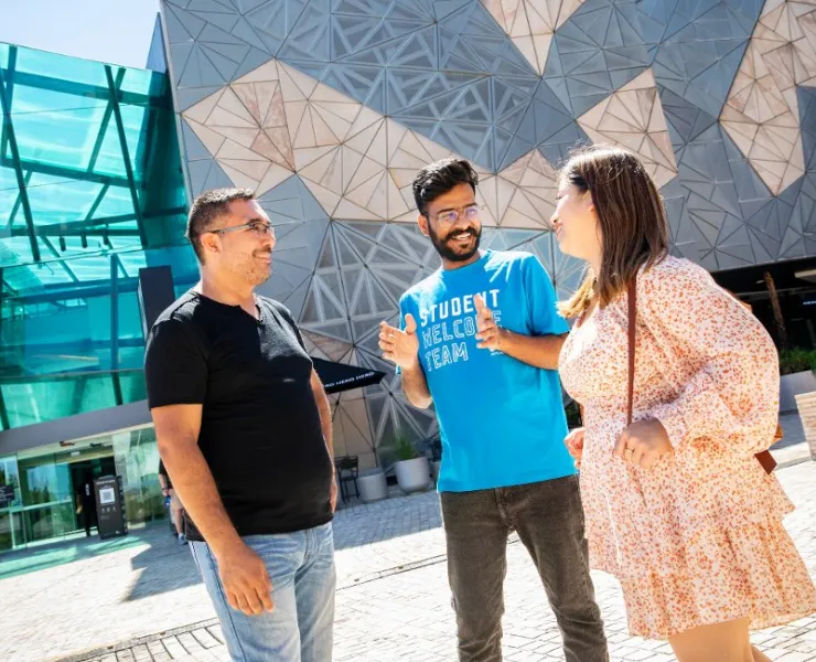 A person in a blue "student welcome team" t-shirt talks to two people in Federation Square.