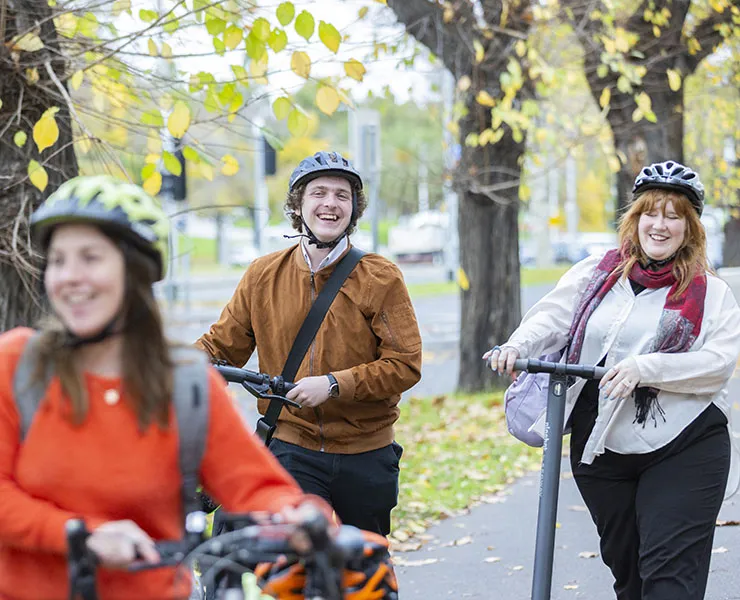 Three smiling people wearing bike helmets, pushing bikes and scooters.