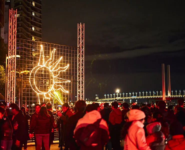Crowd of people at night-time festival with coloured lights, decorations and bridge