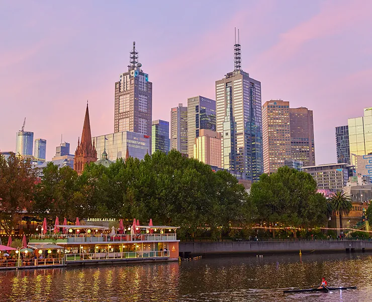 City skyline at disk with purple sky in background and river and boats in foreground.