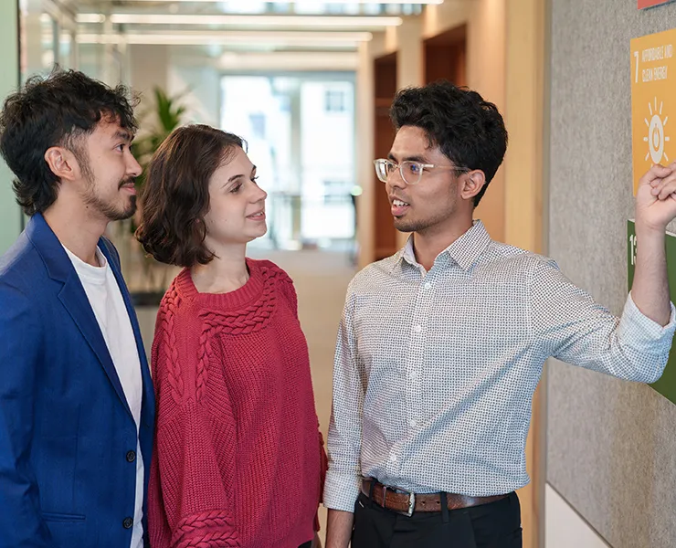 Three people stand talking while one points to a sustainability poster on the wall.