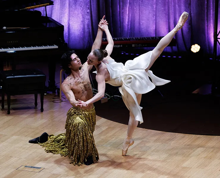  Two dancers pose expressively on stage at Melbourne Fashion Week. One balances on pointe in a white dress while the other, wearing a gold sequined skirt, supports her.