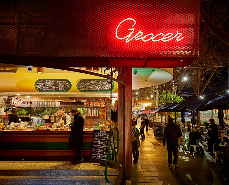 Nighttime scene of a bar in Melbourne with warm lighting, a glowing red neon sign reading ‘Grocer,’ and people walking and sitting at outdoor tables along the footpath.