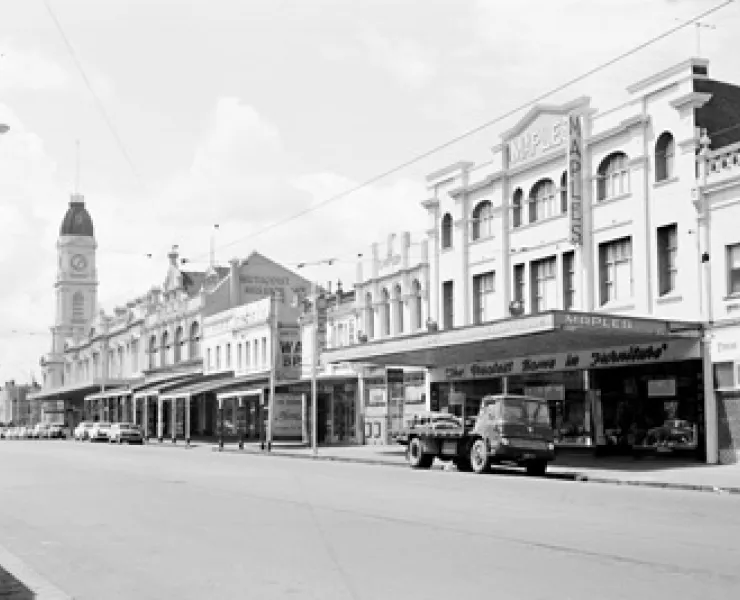 A black and white photograph of a North Melbourne street in the 1960s. Cars are parked outside of businesses.