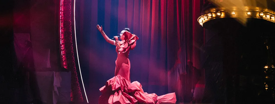 A woman dressed in a long ruffled dress, sings on-stage with her arm outstretched.