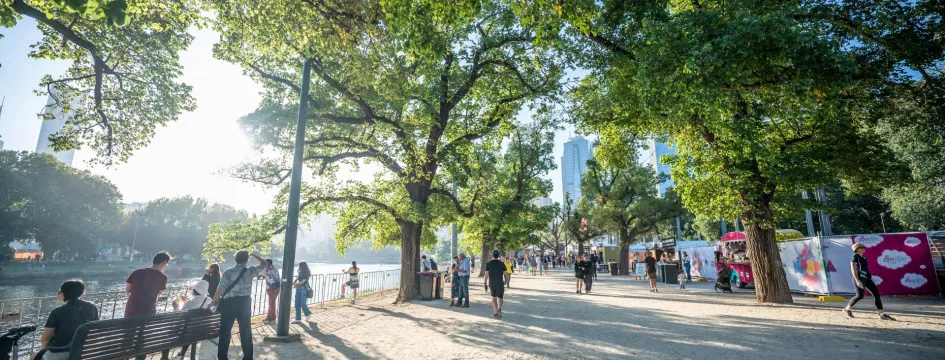 People and trees along a river path in sunlight 