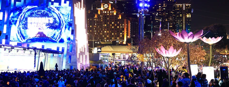 Crowds at an outdoor festival in the city at night. There are lotus flower decorations and a large screen that says 'Buddha's Day'.