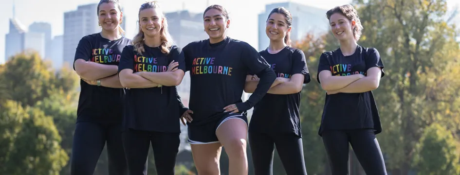 Five women standing in a field with a soccer ball, wearing Active Melbourne tshirts.