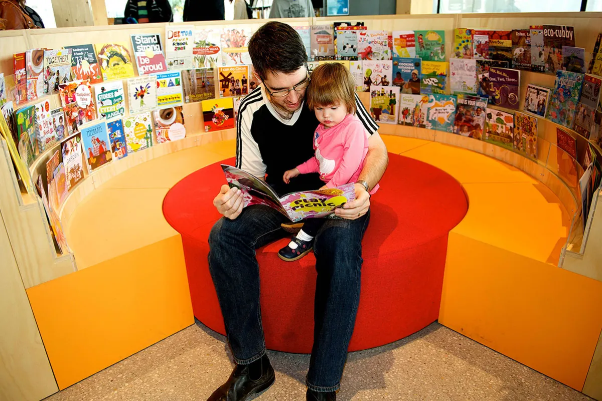 A father reading to his daughter in an enclosed seating area surrounded by children's books.
