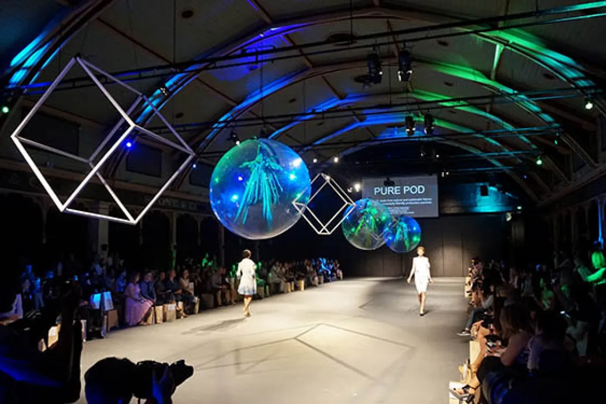 Models on a catwalk under green and blue lights and large shapes suspected from the ceiling.