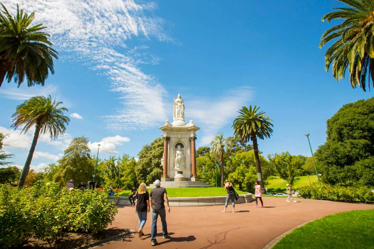 People walking past a monument with a white statue on top and palm trees