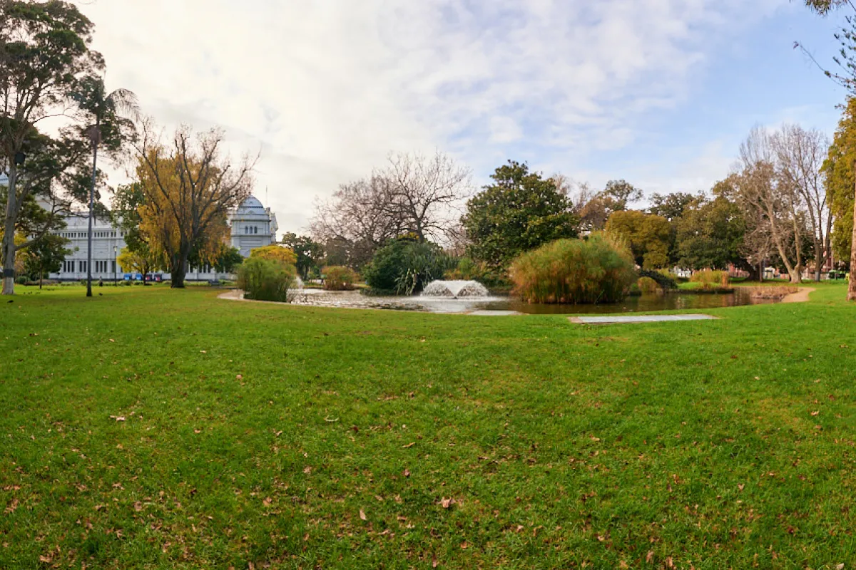 A pond with a fountain surrounded by an expanse of lawn, with trees and the Exhibition Building in the background.