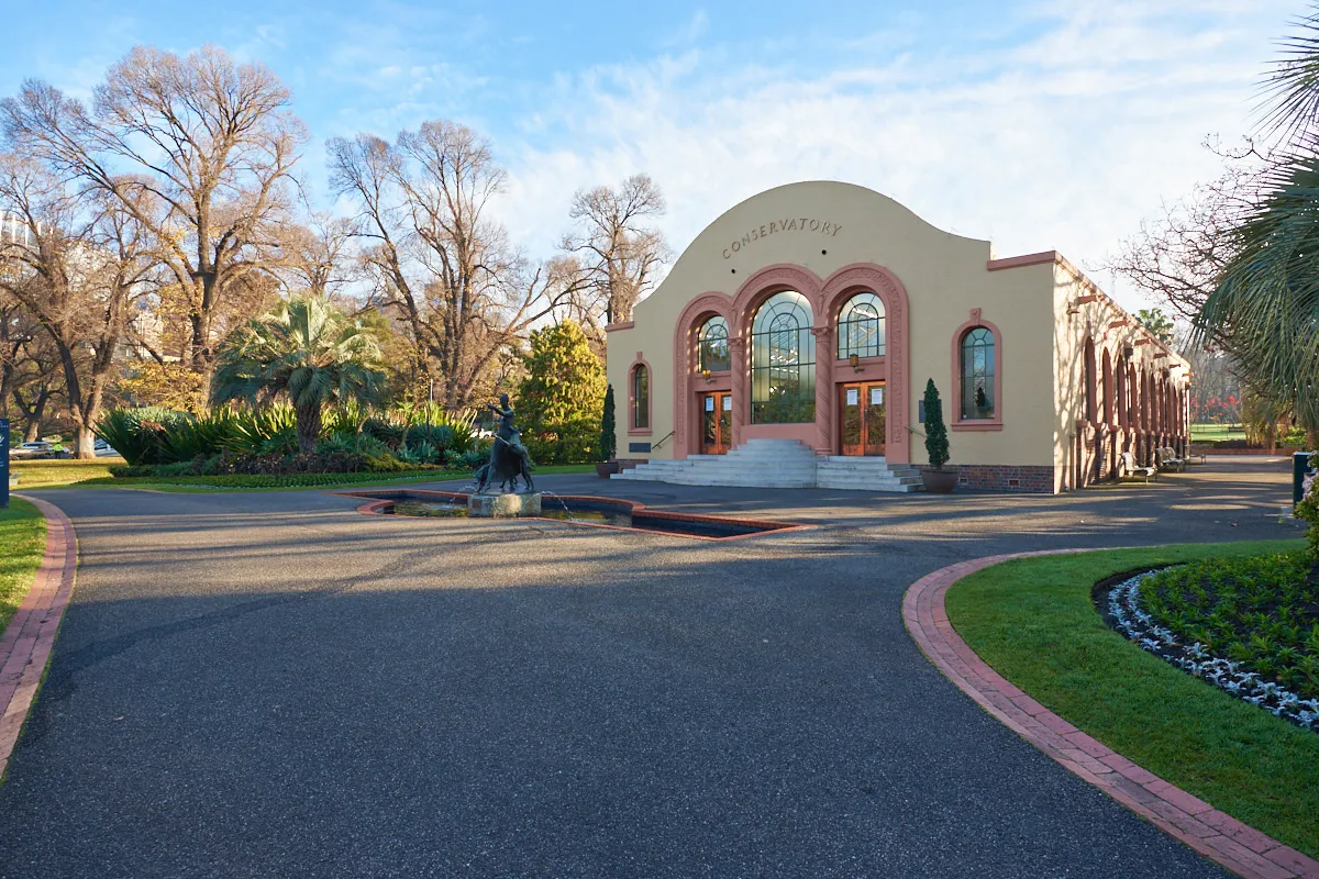 Conservatory building with arched windows and with a statue, fountain and large paved area in front.