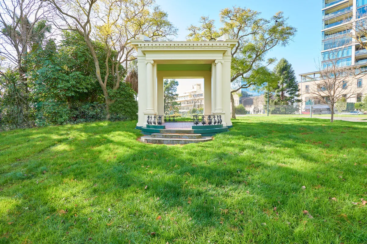 An old bandstand with columns and decorative fencing, surrounded by lawn and with trees and buildings in the background. 
